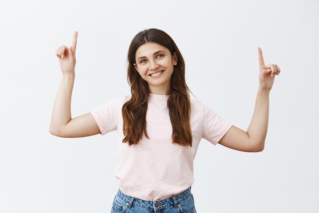 Woman in pink tshirt raising hands pointing upwards and smiling with tilted head