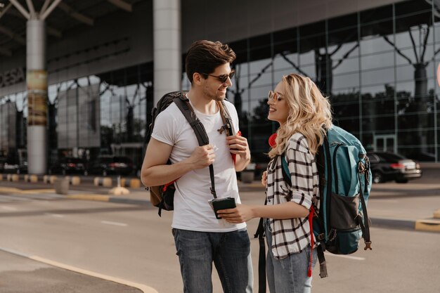 Woman in plaid shirt and man in white tee poses with backpacks near airport Travelers in sunglasses talking and holding passports