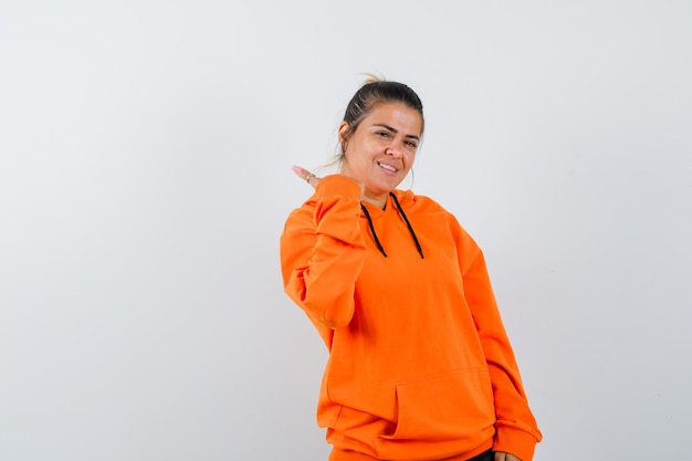 Woman pointing aside with thumb in orange hoodie and looking cheerful