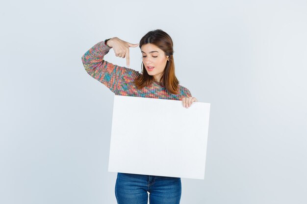 woman pointing at blank board in sweater, jeans and looking confident , front view.