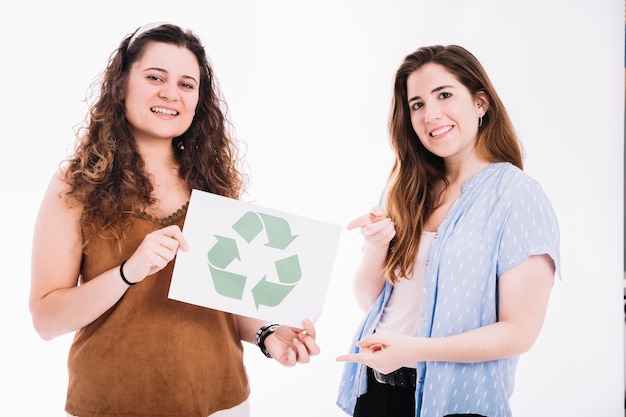 Woman pointing to recycle placard hold by her friend against white backdrop