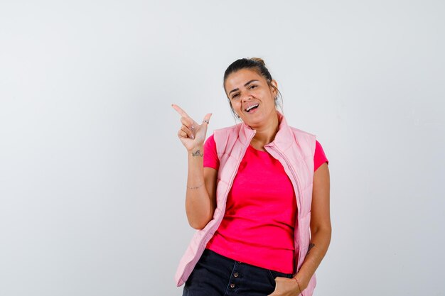 Woman pointing at upper left corner in t-shirt, vest and looking merry 
