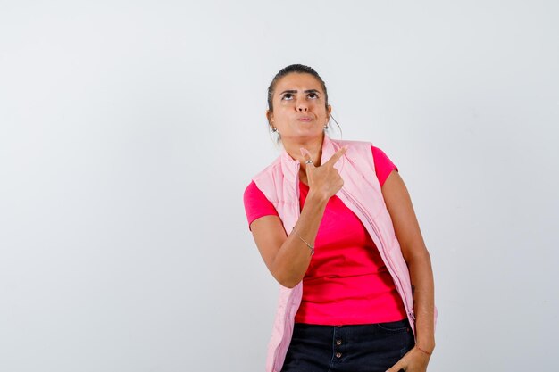 Woman pointing at upper right corner in t-shirt, vest and looking pensive