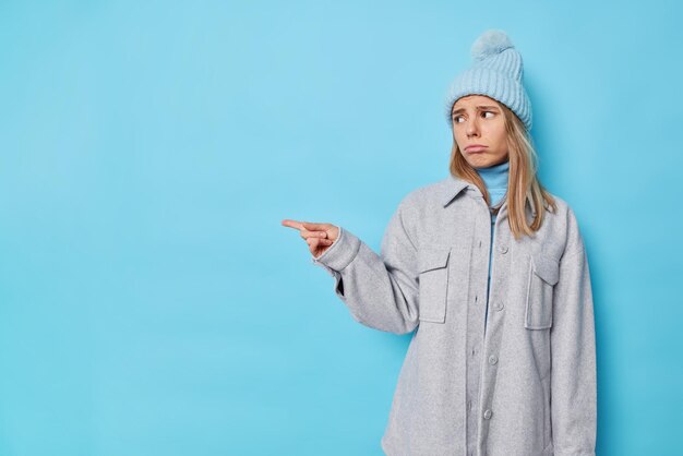 人差し指で女性が指摘する製品が否定的なフィードバックを与える不幸な帽子をかぶって、青で隔離された灰色のジャケットが不満な表情をしています