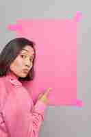 Foto gratuita la donna indica il dito indice al banner di carta rosa vuoto suggerisce di posizionare indossa una giacca rosa attira la tua attenzione su qualcosa.