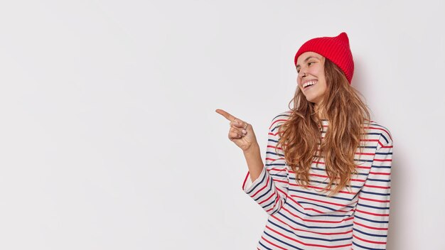 woman points lef at blank copy space demonstrates something awesome incredible thing advertises item wears casual striped jumper and red hat isolated on white.