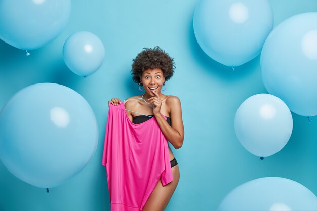 woman poses half naked in lingerie holds pink dress on hangers poses on blue