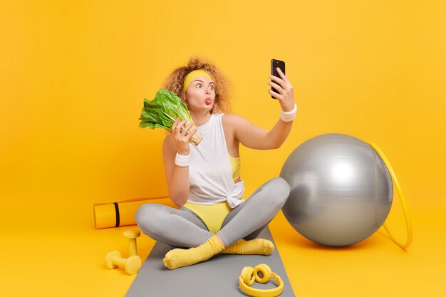 woman poses for making selfie keeps lips folded holds mobile phone eats healthy vegetables keeps to diet sits on mat surrounded by sport equipment 