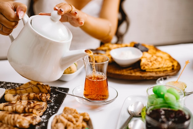 A woman pouring black tea from white ceramic teapot into armudu glass side view