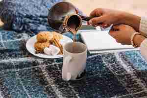 Free photo woman pouring drink in cup from cezve near cookies and notepad