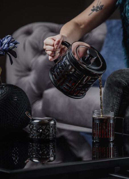 A woman pouring tea from a black teapot into a cup on dark wall side view