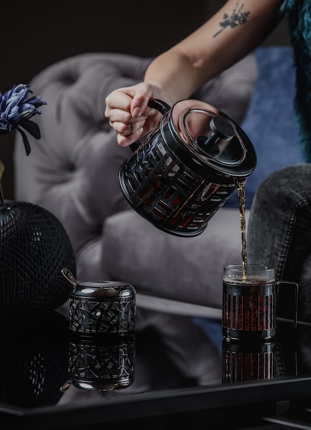 A woman pouring tea from a black teapot into a cup on dark wall side view