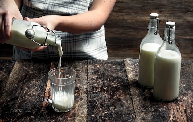 Женщина наливает свежее коровье молоко в стакан. на деревянном столе.