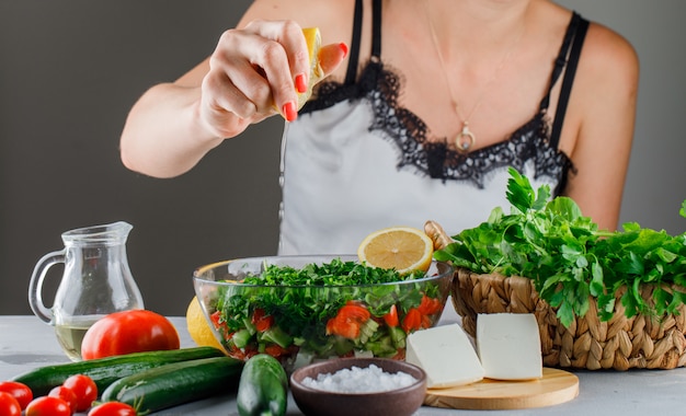 Woman pours lemon juice on salad in a glass bowl with tomatoes, cheese, greens, cucumber side view on a gray surface