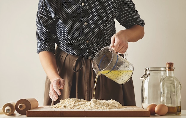 女性は、パスタや餃子の生地を準備するために、ボード上のメジャーカップから小麦粉にオリーブオイルを水に注ぎます。料理ガイドのプレゼンテーション