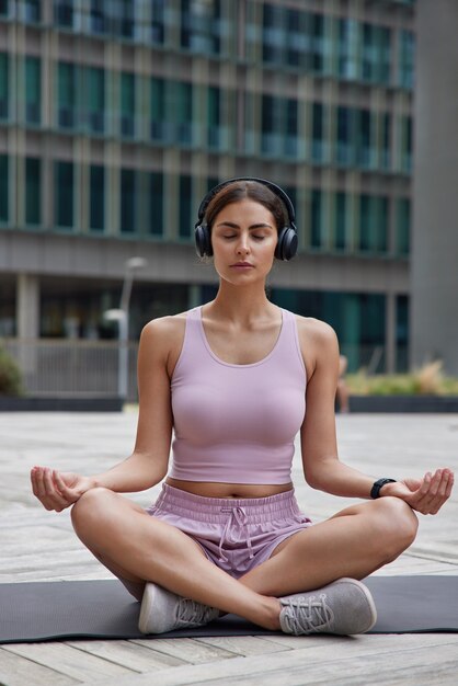  woman practices yoga and meditates concentrated on thoughts during sport training listens music via headphones reaches recreation for mental health poses on fitness mat