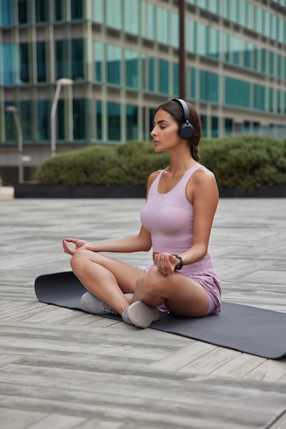  woman practices yoga meditates in harmony outside near office building sits on fitness mat listens music via headphones enjoys relaxation time