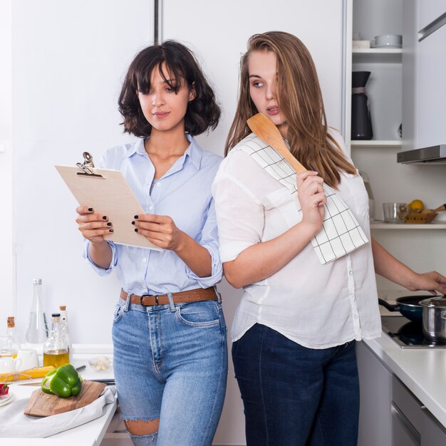 女性が台所でクリップボードにレシピを読んで彼女の友人を見て食べ物を準備します。