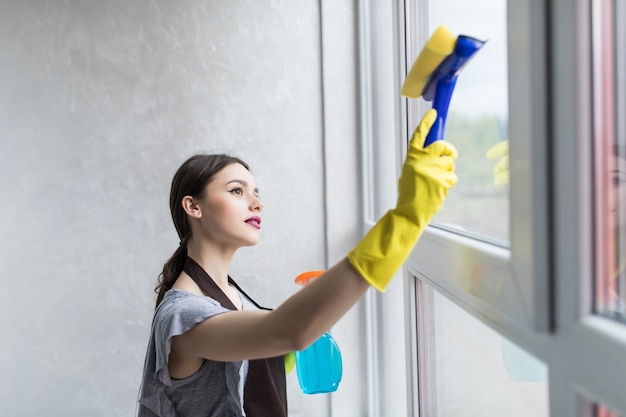 Woman in protective gloves is smiling and wiping dust using a spray and a duster while cleaning her house, close-up