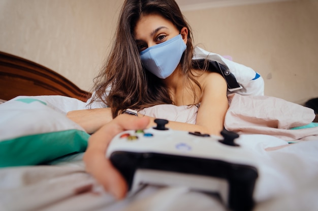 Woman in protective mask lying in bed and holding controller