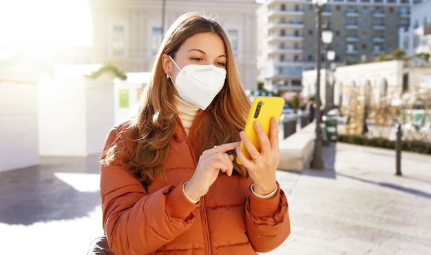 Woman in puffer jacket and medical face mask using her mobile phone outdoors