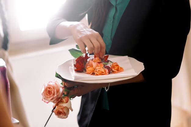 Женщина ставит тонкие апельсиновые розовые бутоны на белую тарелку