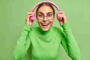 Free photo woman puts on headphones smiles happily listens audio track has glad mood wears round spectacles and turtleneck on green