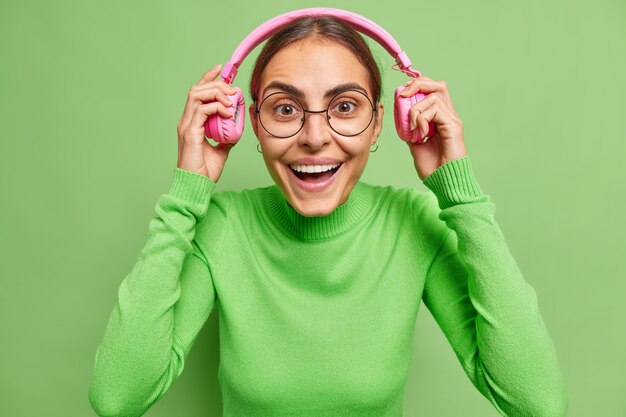 женщина надевает наушники улыбается, радостно слушает аудиозапись, в хорошем настроении носит круглые очки и водолазку на зеленом