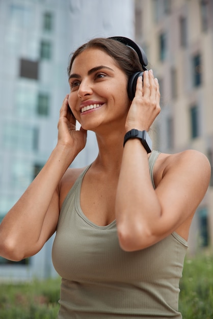 Free photo woman puts on stereo headphones smiles gently dressed in sportswear uses smartwatch for tracking fitness activity poses