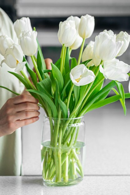 Женщина кладет букет цветов белых тюльпанов в вазу на кухне