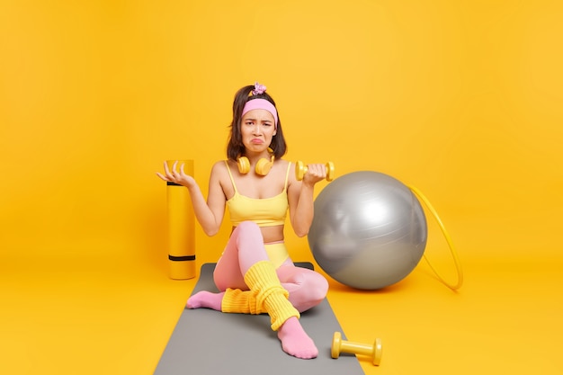 woman raises dumbbell sits on fitness mat dressed in sportswear dos aerobics at home has displeased expression 