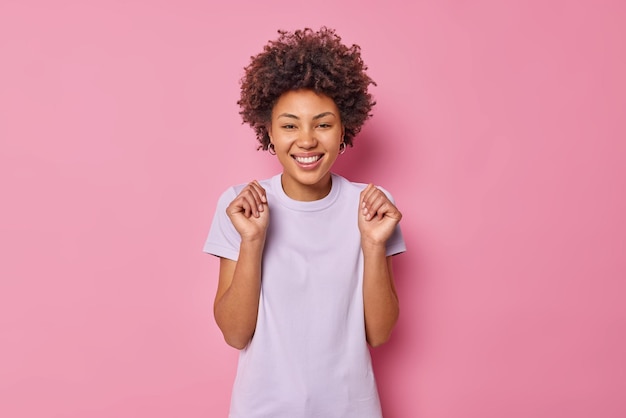 Free photo woman raises fists celebrates success smiles joyfully wears casual t shirt enjoys lucky moment dressed in casual t shirt isolated on pink rejoices victory
