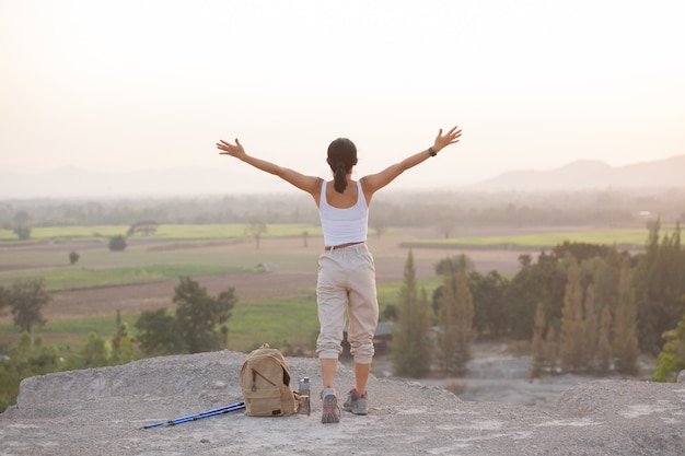 Woman raising hands up on the top of a mountain while hiking and poles standing on a rocky mountain ridge looking out valleys.