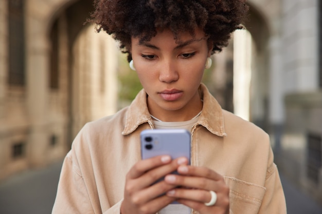 Foto gratuita la donna legge il messaggio sms sullo smartphone naviga in internet senza fili vestita con una giacca marrone casual pone all'esterno usa l'app software