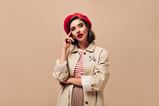 Woman in red beret and coat thoughtfully posing on beige background.  Modern lady with bright lipstick in striped sweater and stylish cloak looks away.