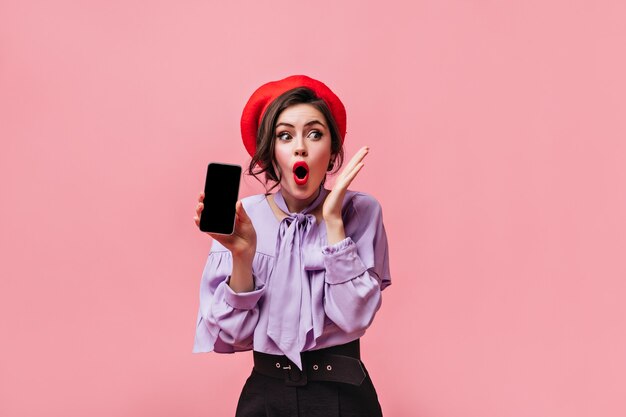 Woman in red hat and lilac blouse is holding black smartphone and posing in shock over pink background.