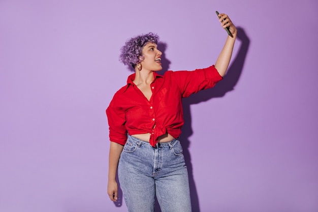 Free photo woman in red long-sleeved shirt and tight-fitting jeans takes selfie on. cute woman with stylish haircut posing.