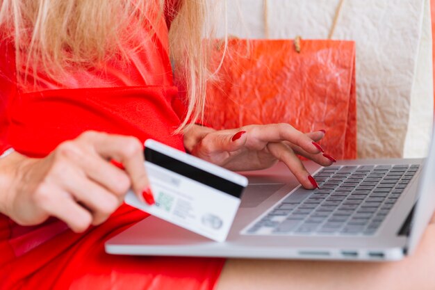 Woman in red sitting with laptop and card near shopping bags 