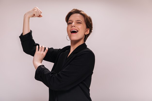 Woman rejoices to her power and demonstrates her biceps. Charming lady in black jacket smiles and shows muscles on isolated background