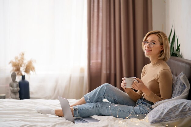 편안 하 고 침실에서 랩톱 컴퓨터를 사용 하여 뜨거운 커피 또는 차 한잔 마시는 여자.