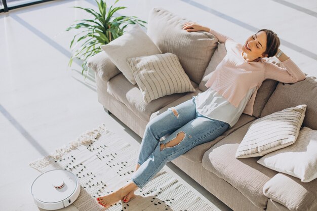 Woman relaxing on sofa while robot vacuum cleaner doing housework