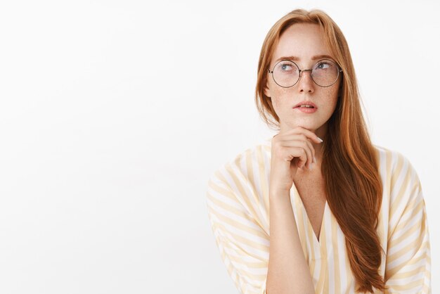 Woman remembering schedule while planning next work day standing focused and perplexed in trendy glasses looking at upper left corner thoughtful and concentrated making calculations in mind