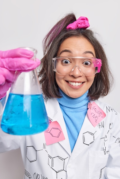 Woman researcher works in chemical laboratory holds beaker with blue liquid does scientific research conducts experiment wears uniform and protective transparent glasses makes new investigation