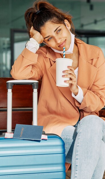 Woman resting on her luggage at the airport and having coffee during pandemic