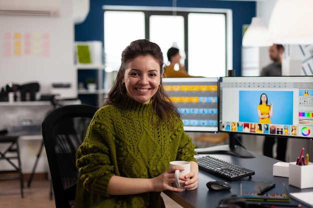 Woman retoucher looking at camera smiling sitting in creative media agency retouching client photos on PC with two displays