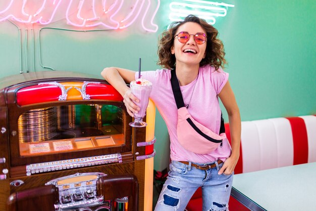 woman in retro vintage 50's cafe at music jukebox drinking milk shake cocktail in hipster outfit having fun laughing in cheerful mood