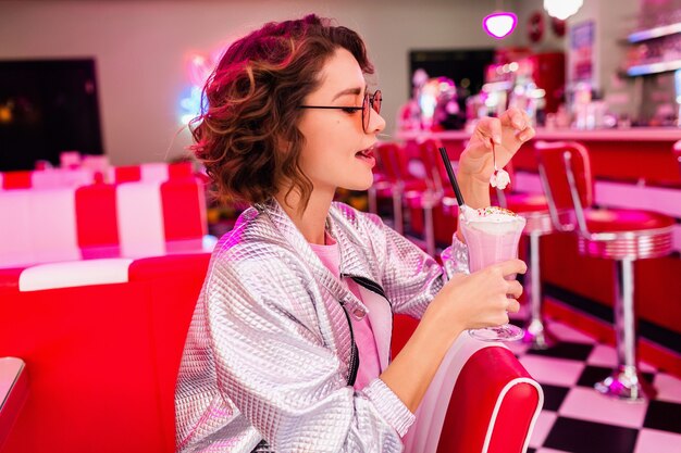 woman in retro vintage 50's cafe of pink color sitting at table drinking milk shake cocktail in hipster outfit having fun