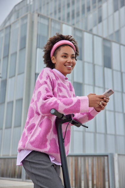 女性が街中の電動スクーターに乗るファッショナブルな服を着た幸せな表情は現代の電気輸送を使用し、オンラインのスマートフォンチャットは新しい場所を発見します