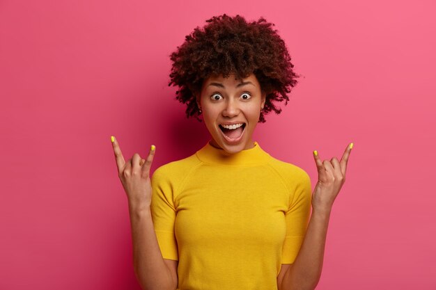 女性のロックンロールスターは、ヘビーメタルのサインを作り、喜びを感じ、目をつぶって口を開け、黄色のTシャツを着て、ポジティブな雰囲気をもたらし、クラブでクールな音楽を楽しんでいます。