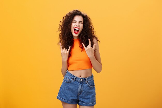 Woman rocking and enjoying great concert dancing yelling out loud from amazement and thrill showing rock-n-roll gesture opening mouth and squinting posing in stylish outfit over orange background.